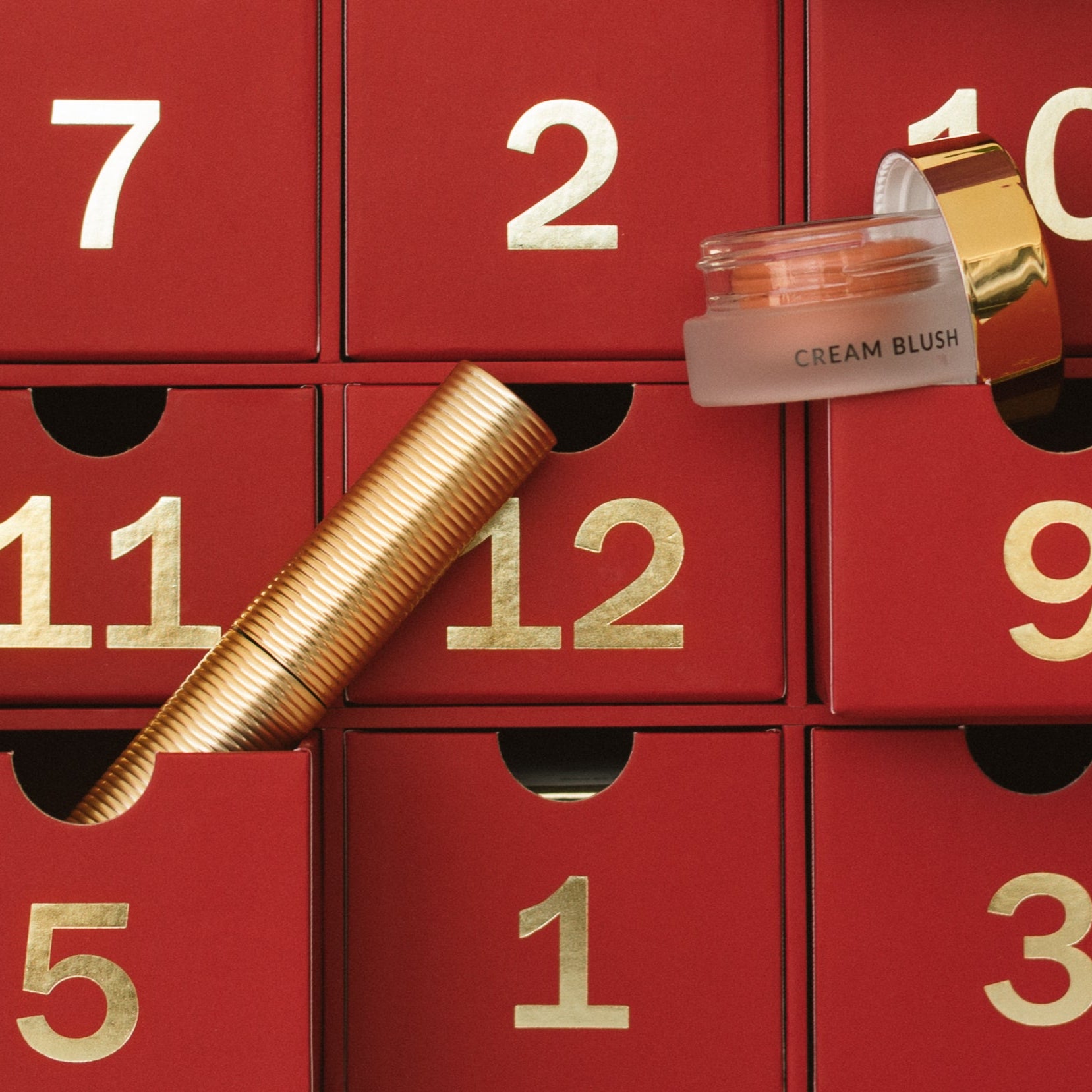 Red advent calendar with gold numbered drawers, featuring a cream blush and gold tube mascara.