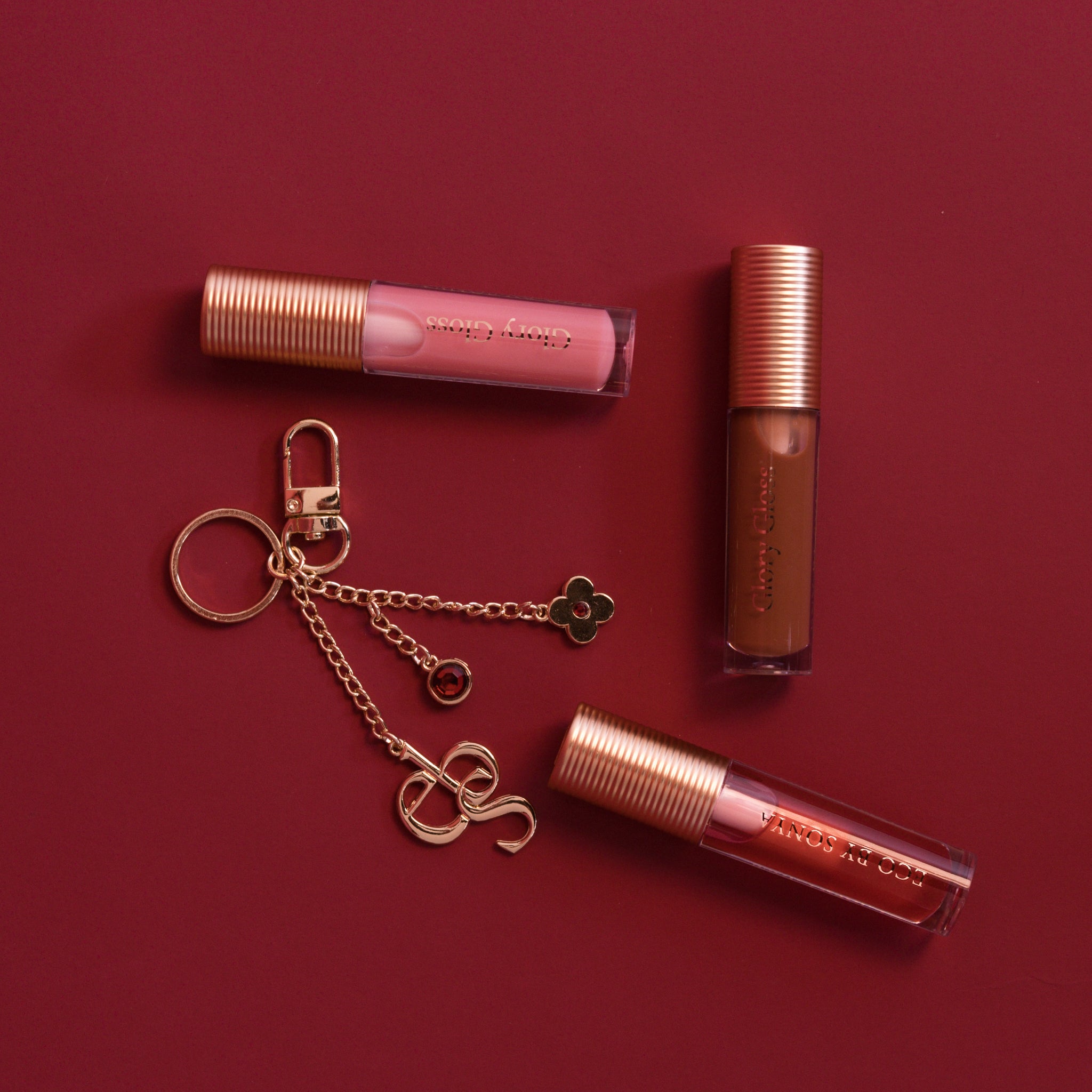Three Glory Gloss shades (Jelly, Candy and Maple) with a gold charm on a deep red background.
