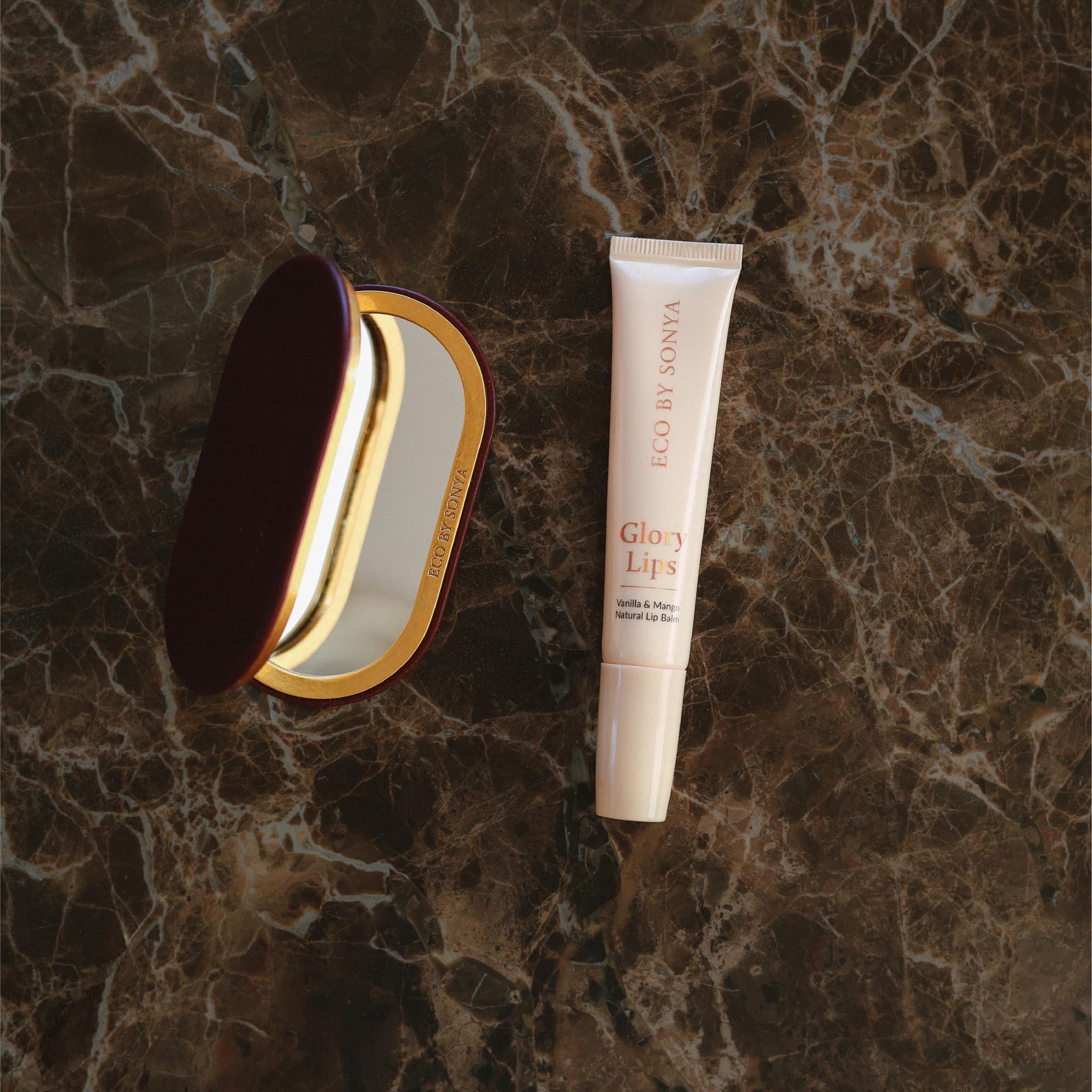 Flat lay of an Eco By Sonya Glory Lips lip balm tube and a mirror styled on a dark brown marble surface.