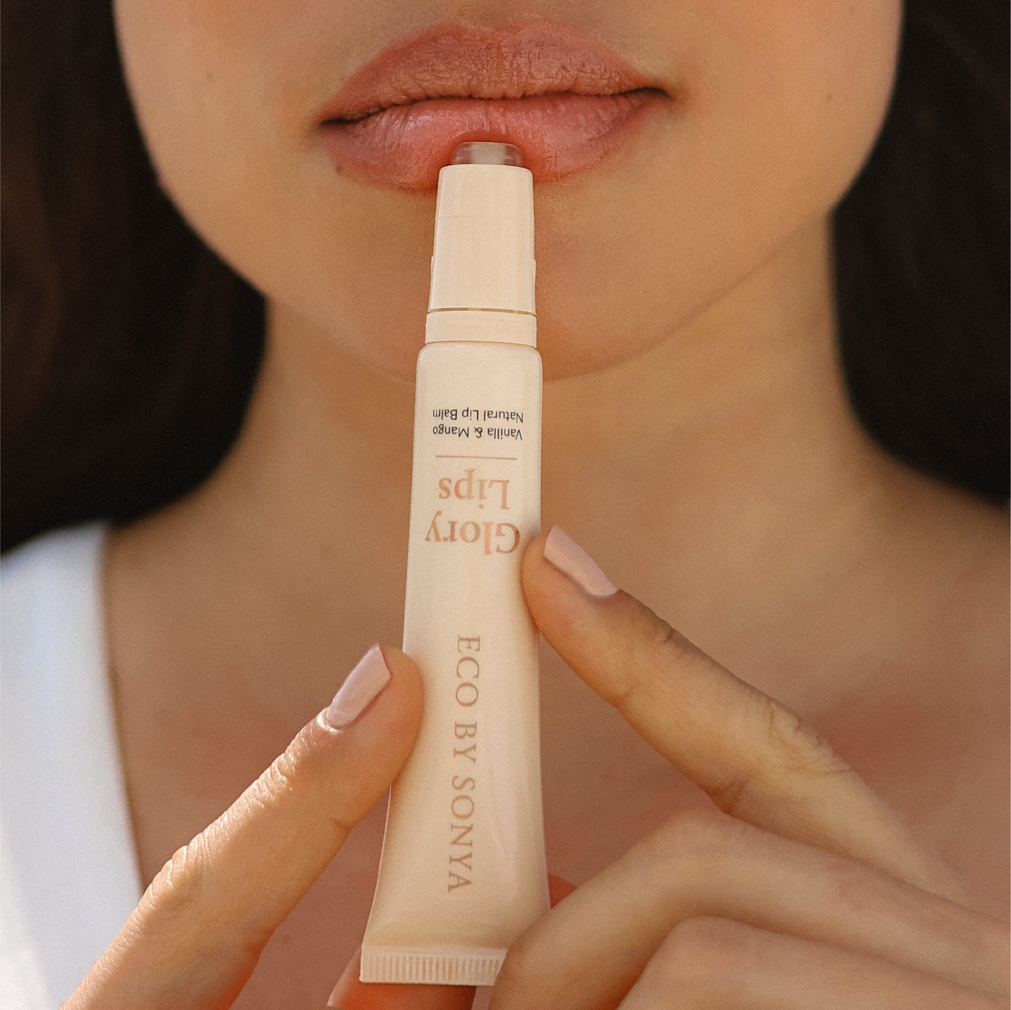Close-up of a person holding an Eco By Sonya Glory Lips Natural Lip Balm against their lips, showing the pale beige tube with gold lettering.