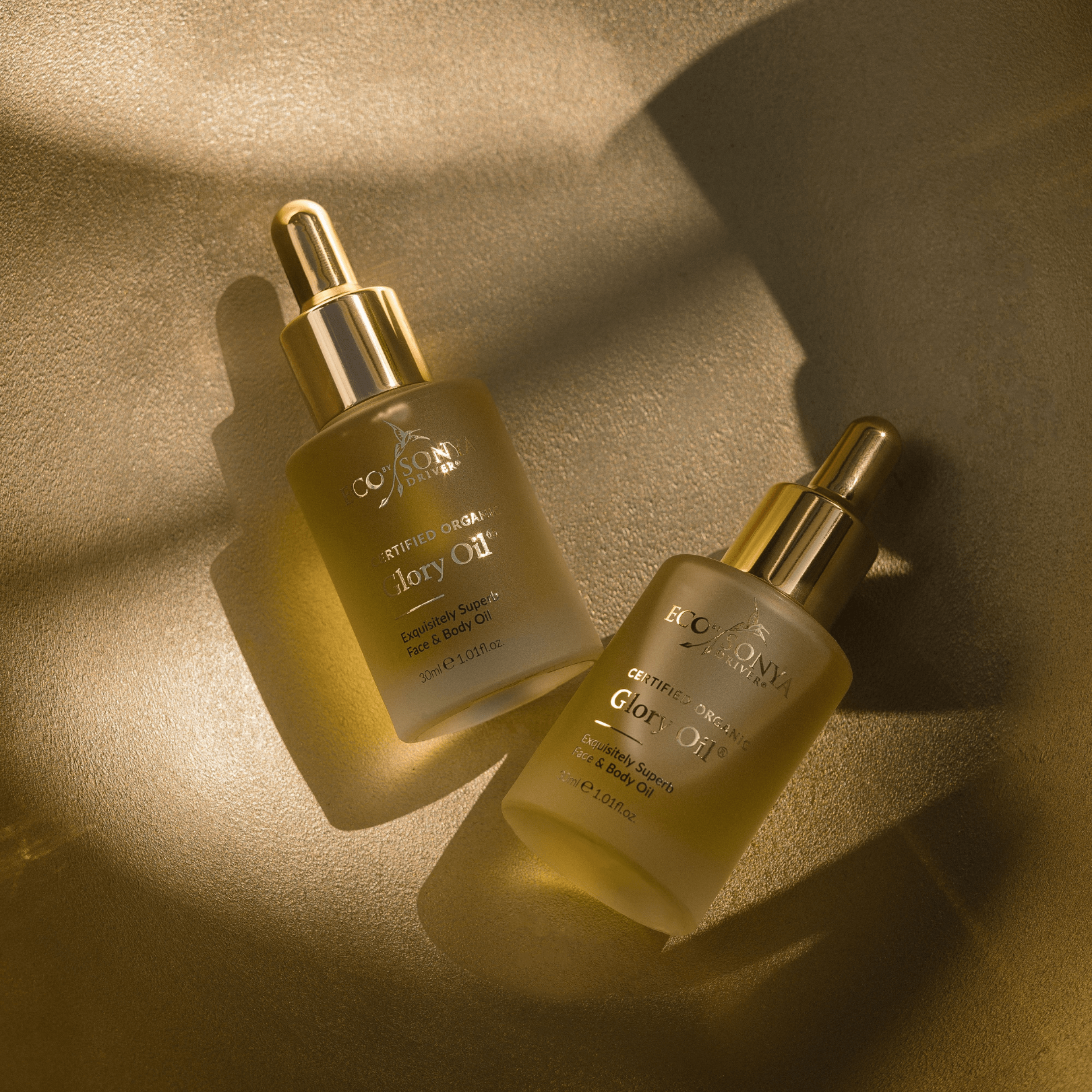 Two Eco By Sonya Glory Oil bottles with golden droppers on golden textured surface, softly lit by sunlight glow.