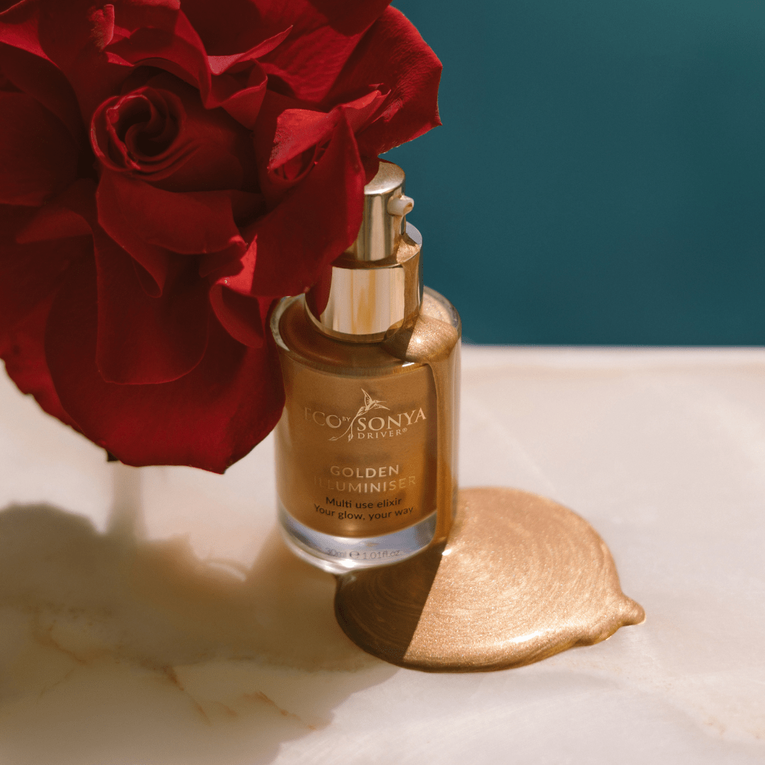 Eco By Sonya Golden Illuminiser bottle with golden product dripping beside red rose on marble surface, glowing liquid texture