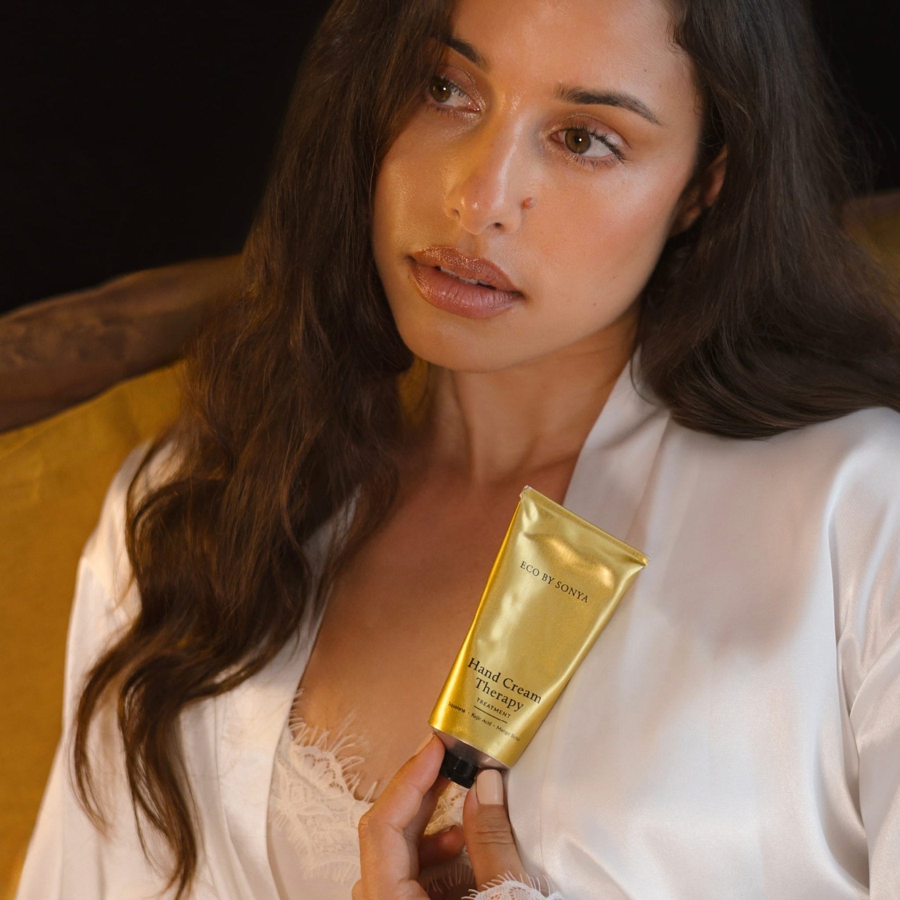 Woman in a white satin robe holding a gold tube of Eco By Sonya Hand Cream Therapy Treatment against a dark background.