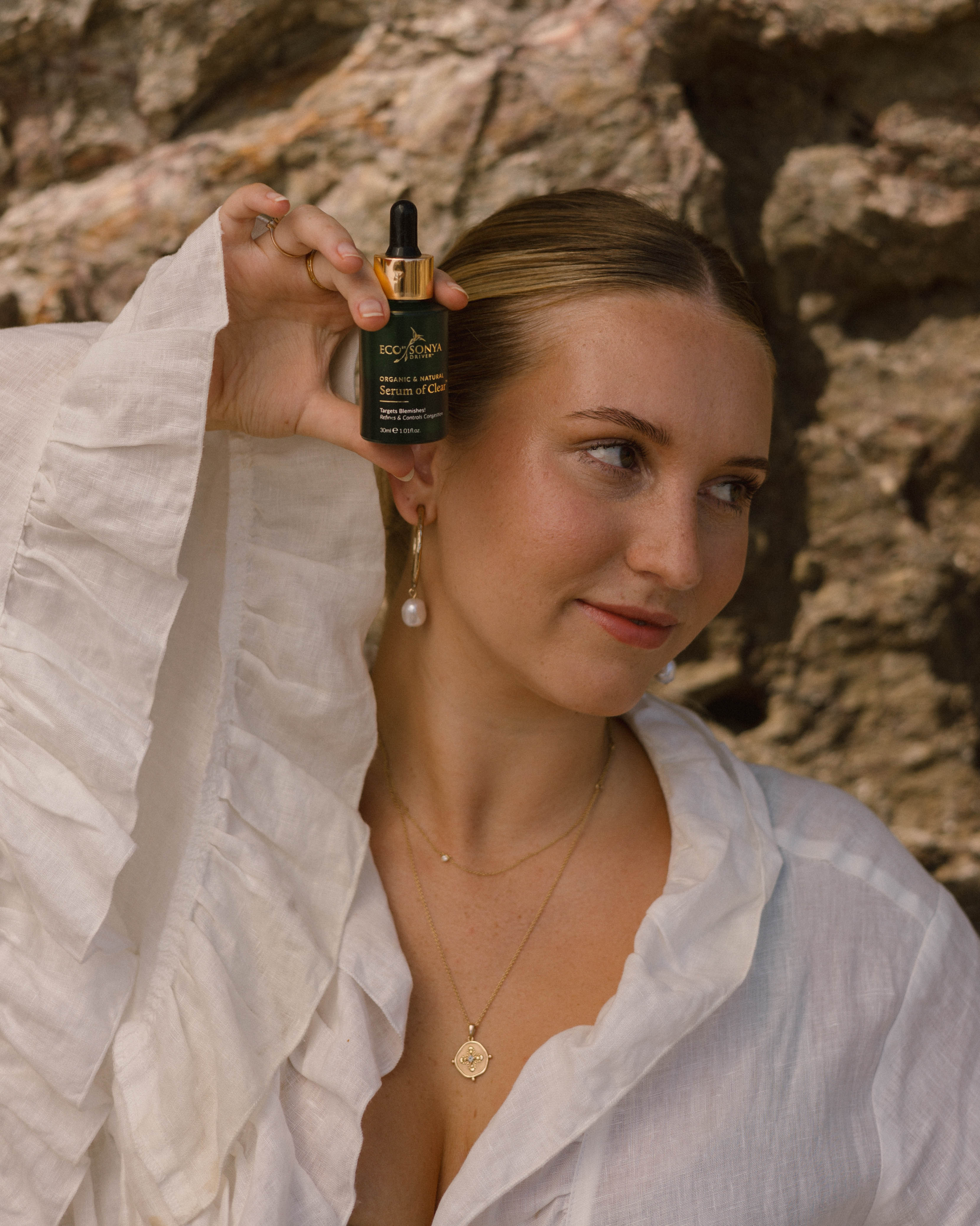 Woman holding a bottle of Eco by Sonya Serum of Clear against a stone wall.