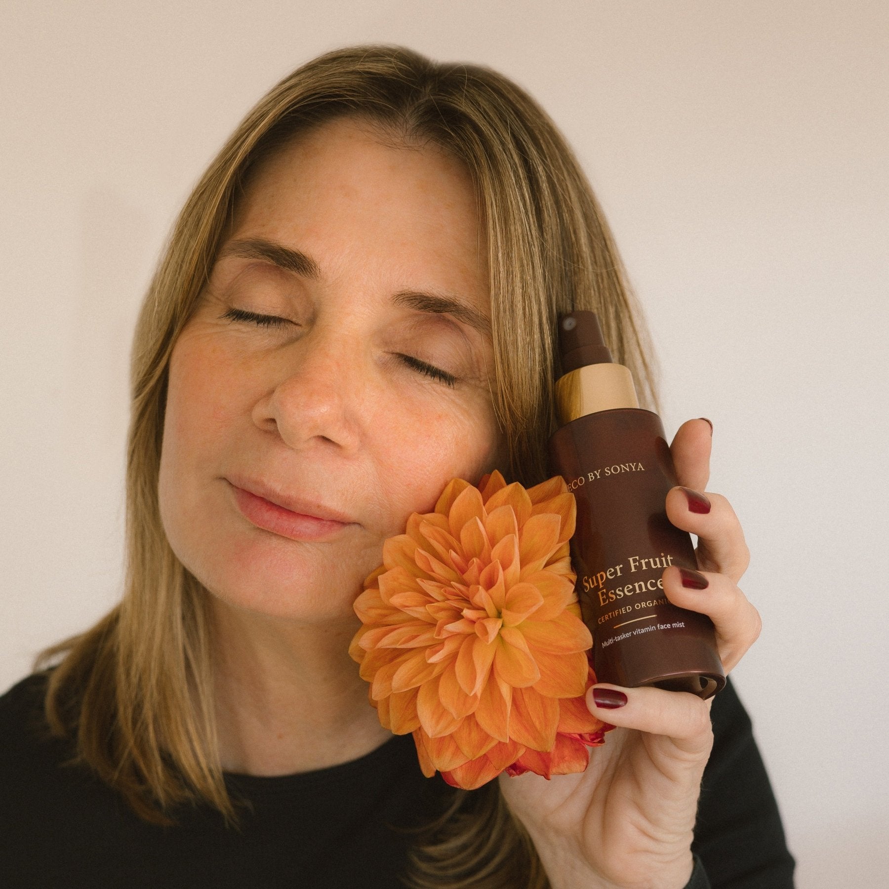 Woman holding an orange flower and a bottle of Eco By Sonya Super Fruit Essence against a plain background