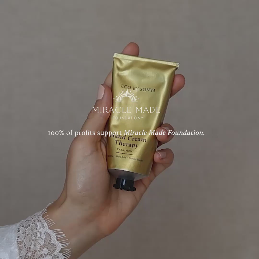 Woman in a white satin robe holding a gold tube of Eco By Sonya Hand Cream Therapy Treatment against a dark background.